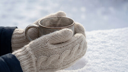 Warm hands in knitted mittens holding a steaming cup of beverage, surrounded by soft white snow, evoking a cozy winter atmosphere and inviting comfort