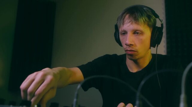 Young male sound designer with headphones working on a mixing console, professionally adjusting faders and knobs to create and produce electronic music in a professional recording studio