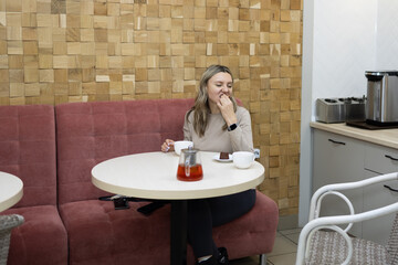 Young woman in a beige shirt sits at a round table in a cafe, eating dessert and drinking tea. She smiles and looks pleased while enjoying her afternoon treat.