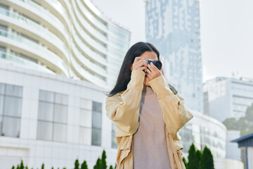 Fototapeta premium Woman with camera adjusting lens for street photography in an urban cityscape, wearing a beige jacket, capturing candid moments among modern buildings and glass towers