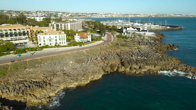 Aerial view of Campos de Lapias in Cascais showing rocky cliffs, winding coastal road, Santa Marta lighthouse and marina with yachts under sunny skies.