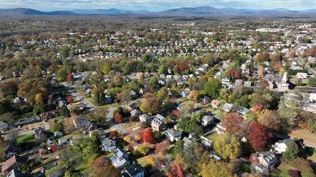 Sweeping drone shot captures a charming suburban neighborhood in the United States during peak autumn, showcasing rows of houses and tree-lined streets bursting with fall colors under a midday sky.