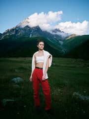 Naklejka premium Woman standing in a grassy field with distant mountains, wearing red pants and a white crop top, cardigan over shoulders, outdoor fashion, rugged landscape.