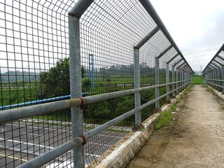 Industrial pedestrian flyover featuring a galvanized steel safety mesh fence and concrete walkway overlooking a green landscape and highway. Secure infrastructure design.