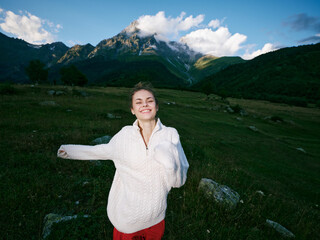 Naklejka premium Smiling woman in a white sweater stands in a green meadow with distant mountains, open sky and rolling hills, capturing freedom, nature, and outdoor exploration in bright daylight.