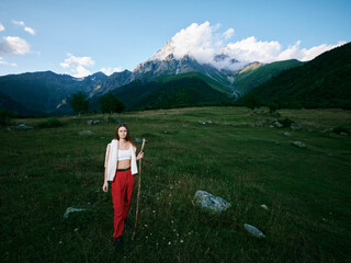 Naklejka premium Woman stands in a green field with a staff, wearing red pants and a white top, mountains in the distance and blue sky behind, depicting outdoor nature and tranquil scenery.