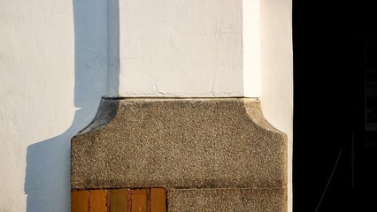 Minimalist architectural close-up of a white plastered wall meeting a textured stone pillar base. Natural sunlight creates sharp geometric shadows on the clean building exterior.