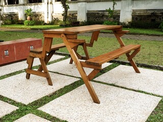 A wooden picnic table and bench set on stone pavers in a peaceful garden. The sunlit outdoor furniture is surrounded by green grass and tropical plants in a relaxing setting.