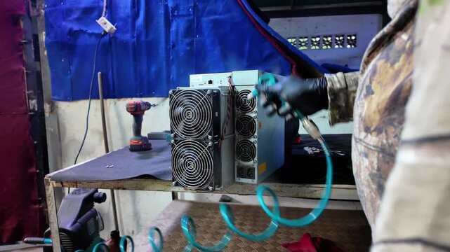 Man using an air hose to clean a cryptocurrency ASIC mining unit. Maintenance tasks on equipment, which is on a workbench next to other power tools.