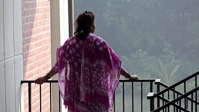 Back view of a Bangladeshi woman in ethnic salwar kameez dress, standing on a high-rise balcony and enjoying the morning outdoor scenery. Lifestyle and leisure in a modern apartment.