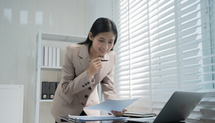 Young Asian businesswoman reviewing printed reports in a bright modern office, smiling confidently while checking data, reflecting professionalism, organization, career growth, corporate success