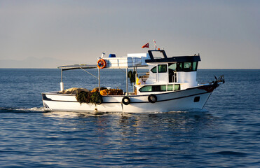 Obraz premium White fishing boat cruising on calm sea—equipped with nets, ropes, lifebuoy, and Turkish flag, framed by distant landforms and clear sky in serene maritime composition.