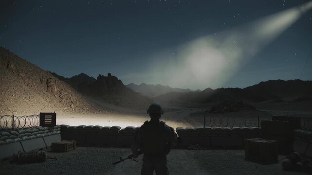 Soldier standing guard in desert outpost under night sky with beam of light illuminating the area for military security