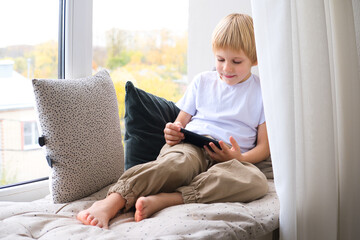 blonde little boy using smartphone sitting on the windowsill