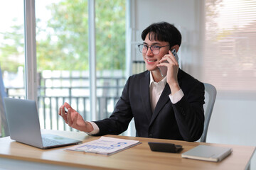 Smiling professional man talking on mobile phone while working on laptop and reviewing documents in a modern workspace