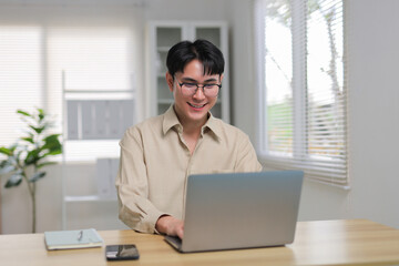 Young Asian man wearing glasses smiling and working on a laptop at a desk in a contemporary, bright office setting
