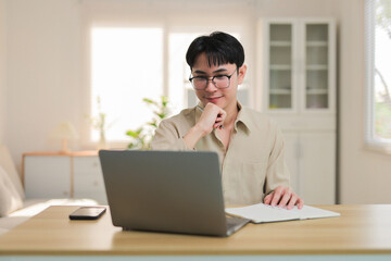 Young Asian man wearing glasses concentrating on laptop, taking notes in a notebook at his bright home office desk