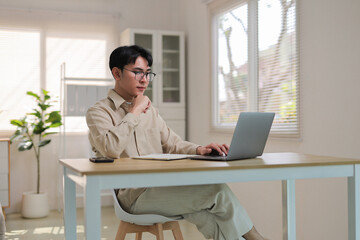 Young Asian man concentrating while working on a laptop at a home office, embodying remote work and freelance lifestyle