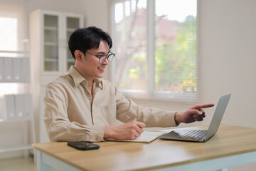 Happy man in glasses at home office laptop during online meeting, smiling while taking notes and explaining on video call