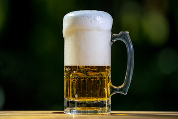 Golden craft beer poured outdoors. Beer splash against green nature background. Pouring amber ale in nature. Brewed beer with bubbles and foam. Craft brewery lager beer on summer day.