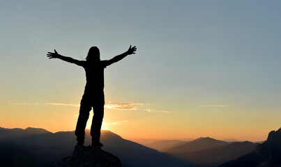 Fototapeta premium Silhouette of a person standing on a mountain with arms raised at sunset, expressing freedom, happiness, mindfulness, and inner peace. Ideal for lifestyle, wellness, and inspirational concepts.