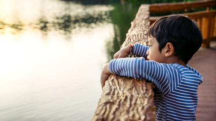 Thoughtful Asian Toddler Leaning on Wooden Railing by Lake, Calm Childhood Moment in Nature