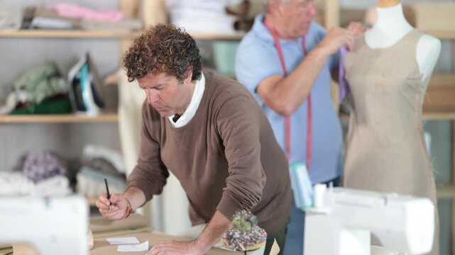  Adult man tailor drawing pattern on paper elderly man assistant sews on mannequin in workshop