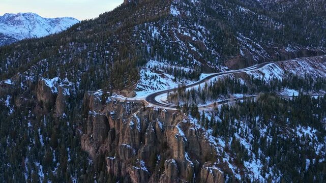 High-angle drone shot of cars driving around a tight hairpin curve on a snowy mountain pass. The winding road cuts through a dense pine forest, highlighting the challenges of winter mountain travel.