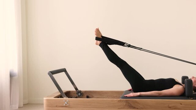 Caucasian woman exercises on a Pilates reformer machine performing leg stretches lying down with straps engaged, promoting flexibility, body balance, strength, and overall fitness concept