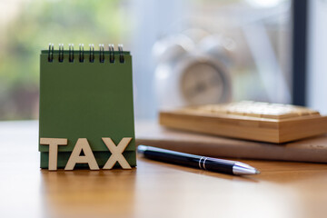 Green flip desk calendar with wooden TAX letters, pen, notebook, and blurred clock in background, symbolizing tax time reminder, financial planning business organization.