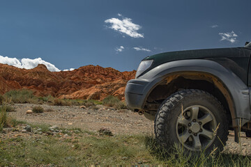An off-road vehicle with aggressive mud tires is parked on a Mongolian dirt road. Medium side-angle shot focuses on the tire tread against a Martian-like landscape of red hills