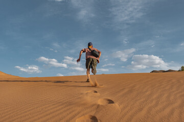 A runner in shorts dashes up a sand dune in the Gobi Desert, leaving footprints behind. Low rear-angle shot captures the sole of his shoe against the vast slope.