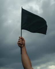 : A hand raises a plain black flag on a thin pole against a backdrop of dark, cloudy skies. The gloomy weather contributes to a somber or ominous atmosphere surrounding the symbol. 