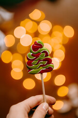 Hand holding Christmas tree shaped candy on stick with warm bokeh lights. Playful festive treat and joyful holiday mood.