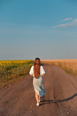A young woman with long brown hair walks along a dirt road, bordered by a field of sunflowers and a grain field.