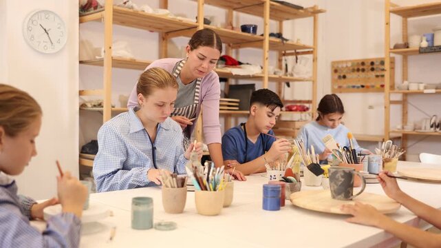 Children and teenagers sit at a table and paint ceramics at the workshop. Teacher shows how to paint a clay product after firing