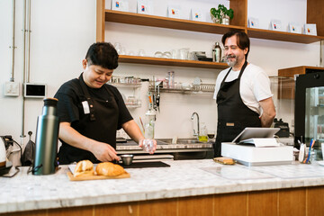 Two baristas, Asian and Caucasian, prepare coffee and pastries behind a cozy cafe counter, surrounded by shelves and kitchenware in a warm, modern coffee shop environment.