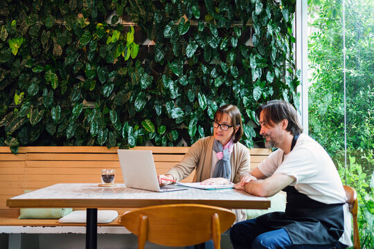 Two colleagues, cafe owner and financial advisor, in a cafe with a laptop discuss work beside a lush indoor green wall, suggesting consulting or small business planning in a modern, casual workspace.