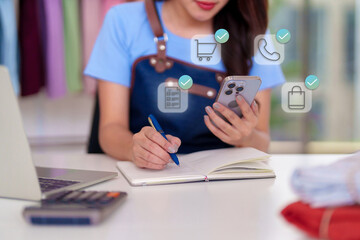 Asian adult woman managing online business orders using smartphone laptop writing notes for ecommerce retail shop small business owner digital entrepreneur work from home