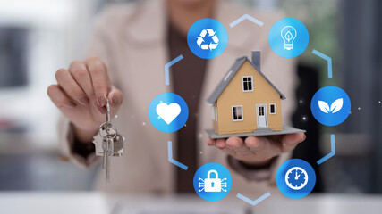 Adult Asian hand holding house keys miniature home model surrounded by smart living technology green energy security icons property concept