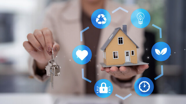Adult Asian hand holding house keys miniature home model surrounded by smart living technology green energy security icons property concept