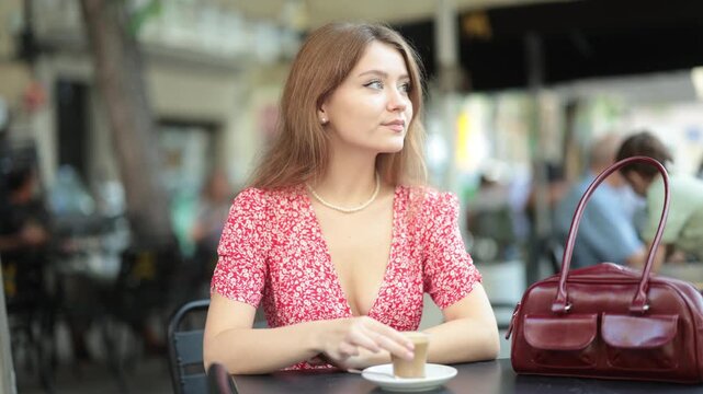 Young woman is sitting at a table in a cafe in a summer dress and drinking Spanish coffee from a glass. Woman tasting cortado coffee with milk. High quality 4k footage