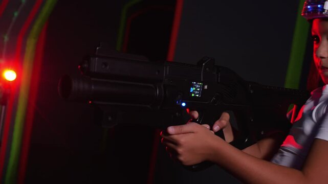 Portrait of an active teenage girl with a laser weapon and headset in a play area. A dark laser tag arena with colorful neon beams on the walls.