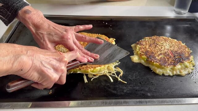 Elderly chef pressing okonomiyaki on the hot teppan in a small restaurant in japan, ensuring the pancakes crisp evenly as they continue cooking side by side