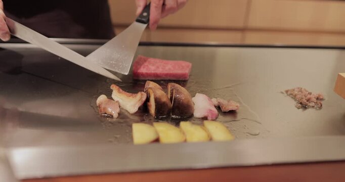 Japanese chef at teppanyaki grill searing marbled Kobe A5 beef, turning the cut then dividing it and setting the pieces on their edge as nearby vegetables continue to sizzle