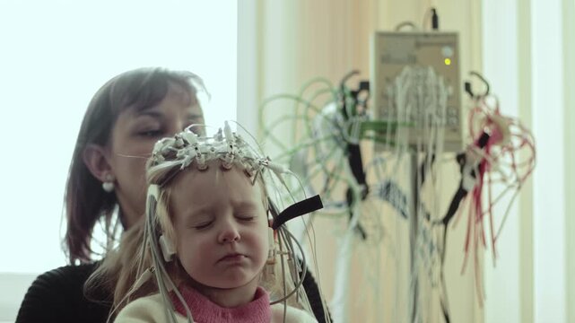 Child undergoing electroencephalography procedure. Electrodes measure spontaneous electrical brain activity. Adult provides support. Medical diagnostic examination. Monitoring equipment indoors.