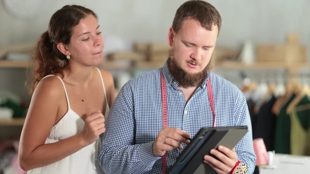 Careful young male dressmaker discussing dress design on tablet with girl in fashion studio . High quality 4k footage