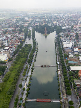 Aerial view of the tranquil river reflects the overcast sky, flanked by lush green trees and the ancient architecture of Hoa Lu, Ninh Binh, Vietnam.