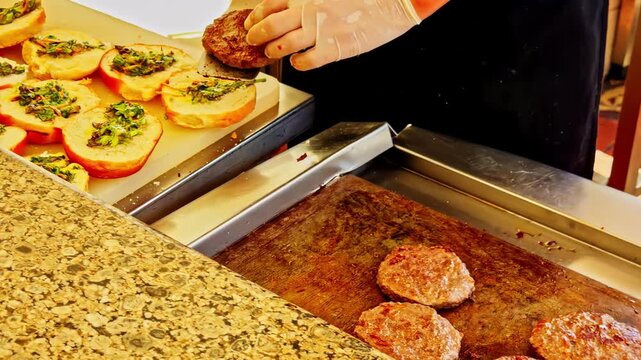 Open buns with herbs and chopped vegetables, and a small burger cooking on the griddle