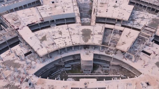 High-angle drone shot over the vast concrete roof of an unfinished commercial complex. Features exposed wiring, open skylight voids, and water stains, symbolizing a halted megaproject.